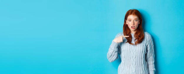 Free photo confused redhead girl pointing at herself being chosen standing in sweater against blue background