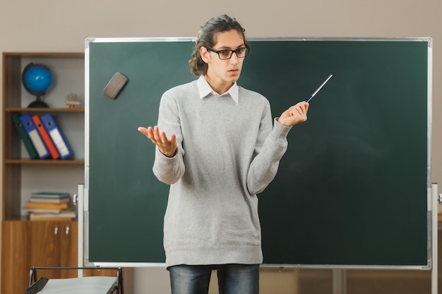 confused spreading hand young male teacher standing on front of blackboard and points at blackboard with pointer in classroom