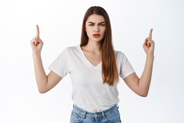 Confused stylish woman frowning pointing up at something strange stare displeased and puzzled questioned by promo deal standing in tshirt against white background