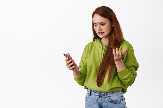 Confused teenage girl looking at smartphone with puzzled face, reading strange message, app error on mobile phone, shrugging questioned, white background