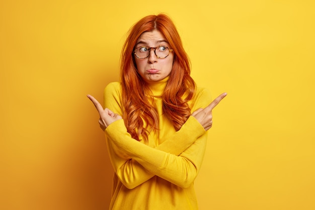 Free photo confused unhappy woman with long ginger hair crosses arms over body and points at different sides hesitates what better to choose wears yellow jumper