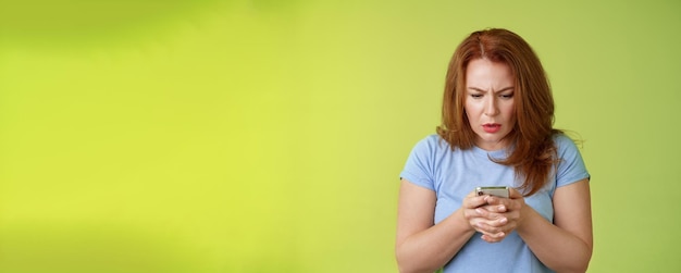 Free Photo confused unsure redhead middleaged woman learn how use social media trying understand emoji look intense focused smartphone display reading important news message stand green background