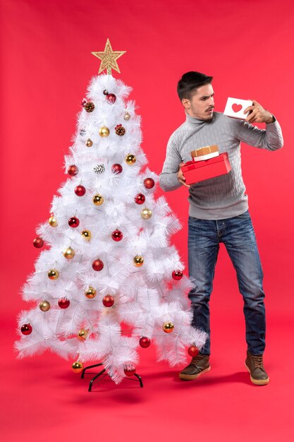 Confused young adult in a gray blouse standing near the decorated Xsmas tree and holding his gifts on red