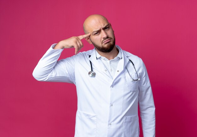 Confused young bald male doctor wearing medical robe and stethoscope putting finger on forehead isolated on pink wall