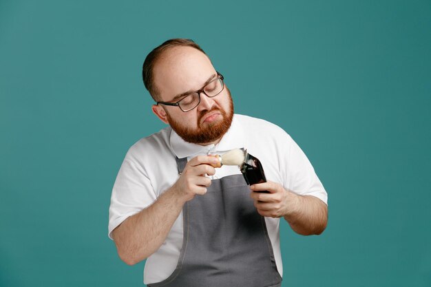 Confused young barber wearing uniform and glasses cleaning hair trimmer with shaving brush isolated on blue background