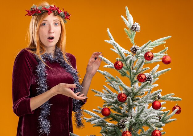 Confused young beautiful girl standing nearby christmas tree wearing red dress and wreath with garland on neck isolated on orange background