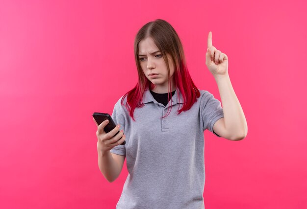 Confused young beautiful woman wearing gray t-shirt looking at phone in her hand points finger to up on isolated pink wall