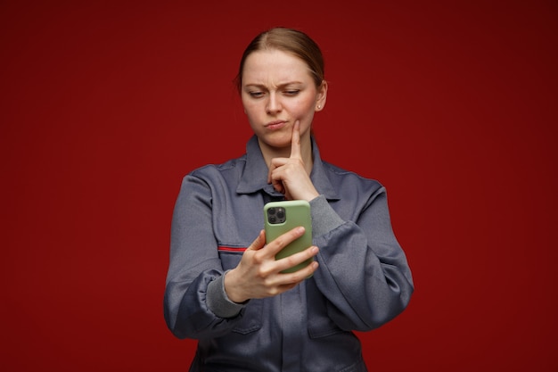 Free Photo confused young blonde female engineer wearing uniform holding and looking at mobile phone touching face with finger 