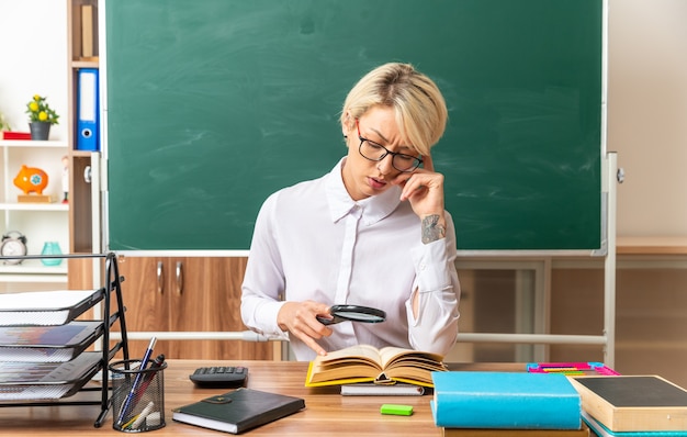 confused young blonde female teacher wearing glasses sitting at desk with school tools in classroom looking at open book through magnifying glass touching head