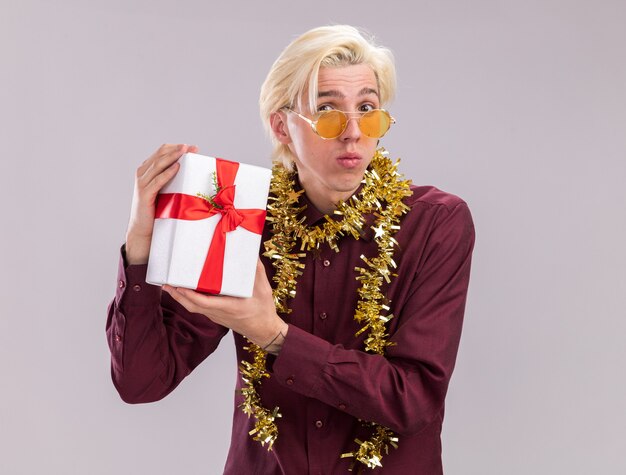 Confused young blonde man wearing glasses with tinsel garland around neck holding gift package looking at camera isolated on white background