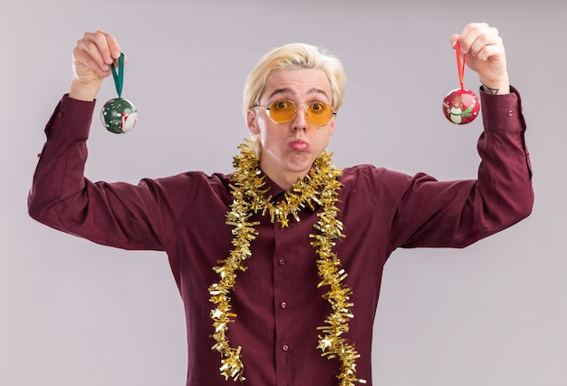 Confused young blonde man wearing glasses with tinsel garland around neck raising christmas baubles up looking at camera isolated on white background