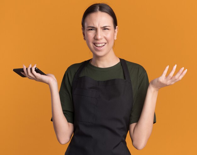 Confused young brunette barber girl in uniform keeps hand open and holds phone on orange