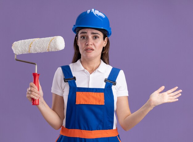 Confused young builder woman in uniform holding roller brush and spreading hand isolated on purple wall