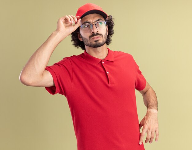 Confused young caucasian delivery man in red uniform and cap wearing glasses grabbing cap looking at side 