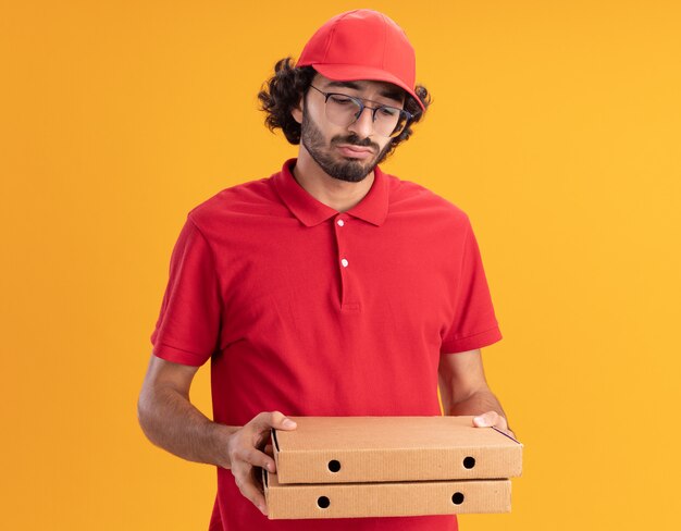 Confused young caucasian delivery man in red uniform and cap wearing glasses holding and looking at pizza packages 