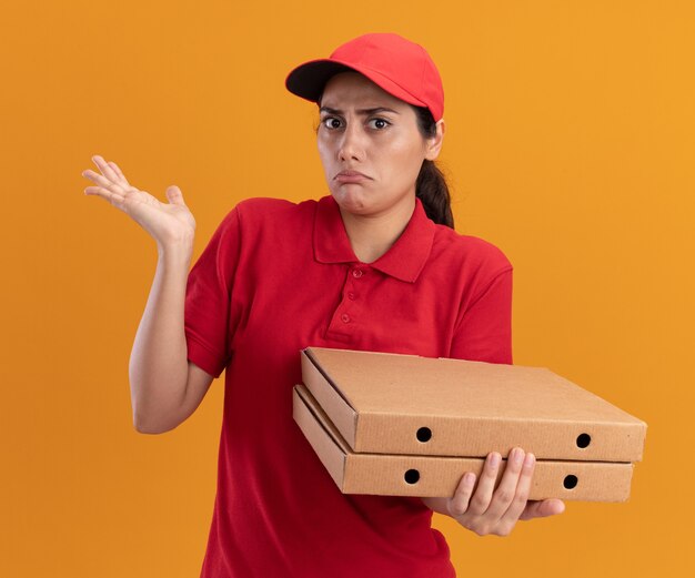 Confused young delivery girl wearing uniform and cap holding pizza boxes spreading hand isolated on orange wall