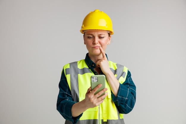 Confused young female construction worker wearing safety helmet and safety vest holding and looking at mobile phone keeping finger on corner of mouth 