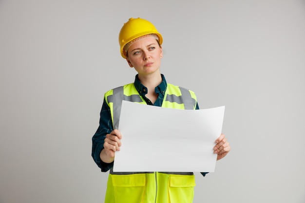 Confused young female construction worker wearing safety helmet and safety vest holding paper looking at side 