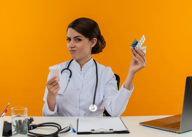 Free photo confused young female doctor wearing medical robe with stethoscope sitting at desk work on computer with medical tools holding empty can and pills on isolation yellow wall