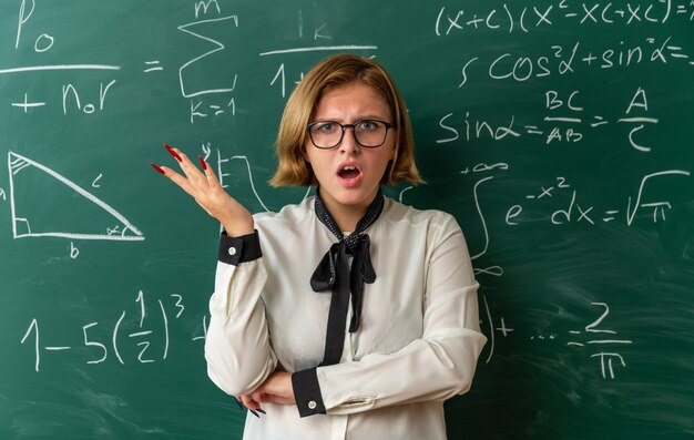 Confused young female teacher wearing glasses standing in front blackboard spreading hand in classroom