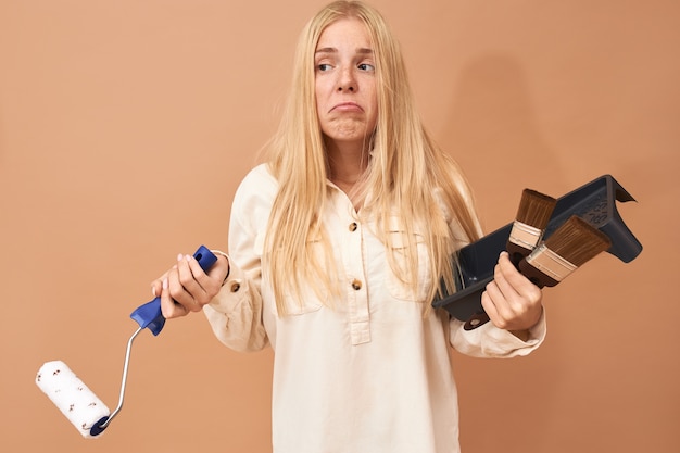 Confused young female with long straight hair posing on blank copy space holding special tools while painting walls