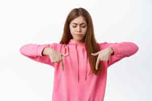 Free photo confused young girl having doubts frowning and pointing down looking with hesitant unsure face standing troubled against white background