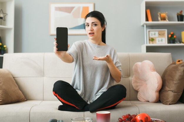 Confused young girl holding and points with hand at phone sitting on sofa behind coffee table in living room