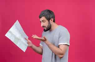 Free photo confused young handsome caucasian traveler man holding and looking at map and showing empty hand isolated on pink  with copy space