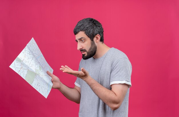 Confused young handsome caucasian traveler man holding and looking at map and showing empty hand isolated on pink  with copy space