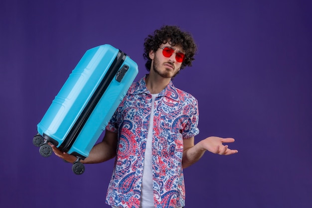 Free Photo confused young handsome curly traveler man wearing sunglasses holding suitcase showing empty hand on isolated purple space with copy space