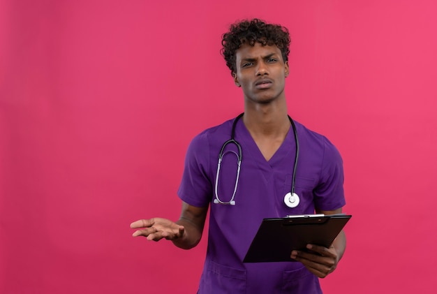 Free photo a confused young handsome dark-skinned doctor with curly hair wearing violet uniform with stethoscope looking angry at camera while holding clipboard