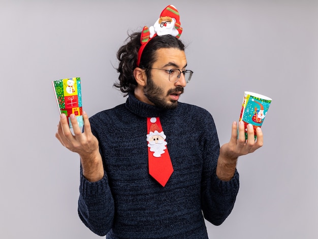 Free photo confused young handsome guy wearing christmas tie with hair hoop holding and looking at christmas cups isolated on white wall