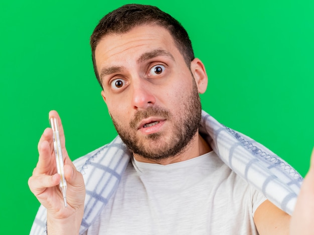 Free photo confused young ill man holding thermometer wrapped in plaid and holding camera isolated on green