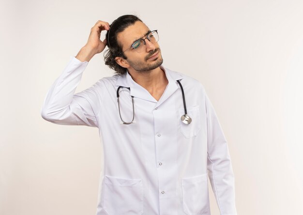 Confused young male doctor with optical glasses wearing white robe with stethoscope putting hand on head