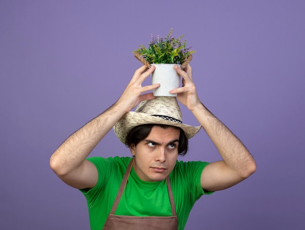 Free Photo confused young male gardener in uniform wearing gardening hat holding flower in flowerpot on head