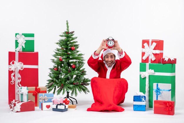 Confused young man celebrate new year or christmas holiday sitting on the ground and holding clock near gifts and decorated xsmas tree checking her time on white background