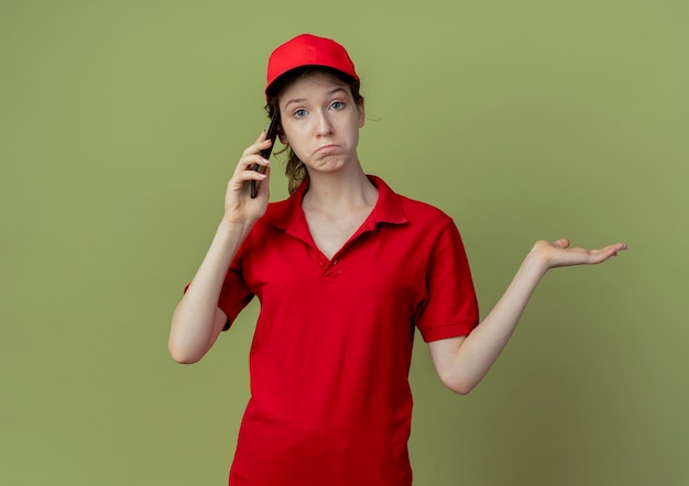 Free photo confused young pretty delivery girl in red uniform and cap talking on phone and showing empty hand isolated on olive green background with copy space