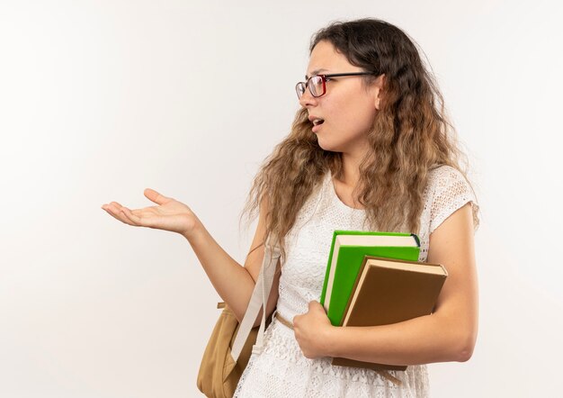 Confused young pretty schoolgirl wearing glasses and back bag holding books looking at side showing empty hand isolated on white 