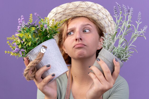 Confused young slavic female gardener wearing gardening hat holding flowers in flowerpots on purple