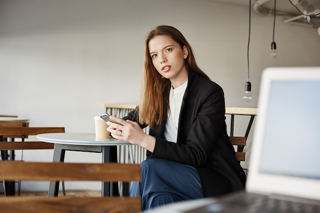 Confused young woman sitting in cafe with smartphone and looking questioned at someone