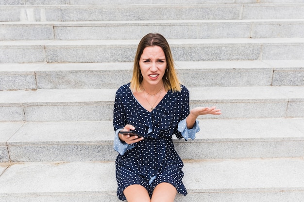 Confused young woman sitting on staircase holding smartphone