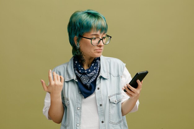 Confused young woman wearing glasses bandana on neck keeping hand in air using mobile phone isolated on olive green background