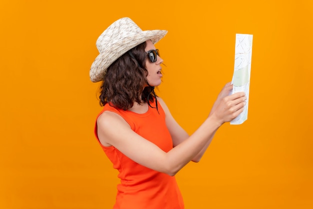 Free Photo a confused young woman with short hair in an orange shirt wearing sun hat and sunglasses raising map with hands and looking attentively at it 