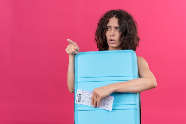 A confused young woman with short hair wearing green crop top pointing with index finger holding blue suitcase with plane tickets