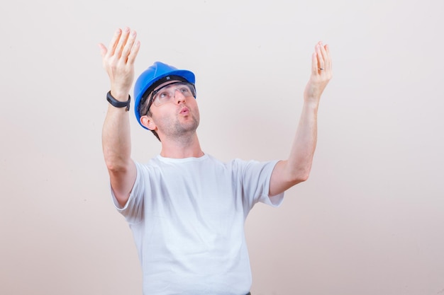 Construction worker raising hands while looking up in t-shirt, helmet and looking surprised