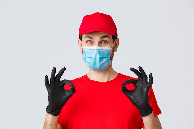 Contactless delivering, covid-19 and shopping concept. Cheerful, pleased courier in red uniform, cap and medical mask with gloves show okay, approval or guarantee gesture, recommend service