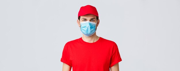 Contactless delivering covid19 and shopping concept Closeup of young man courier in red uniform cap and medical mask looking at camera advertisement of deliver company