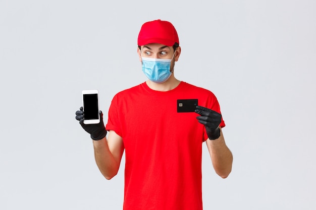 Contactless delivery, payment and online shopping during covid-19, self-quarantine. Excited courier in red uniform, face mask and gloves, look smartphone display, show credit card, paying order.