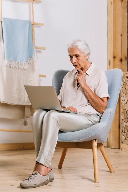 Contemplated senior woman sitting on armchair looking at laptop