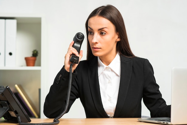 Contemplated young businesswoman sitting in the office holding telephone receiver